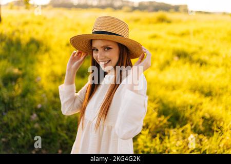 Portrait moyen d'une jeune femme au gingembre, portant un chapeau de paille et une robe blanche debout, posant sur un magnifique champ d'herbe verte Banque D'Images