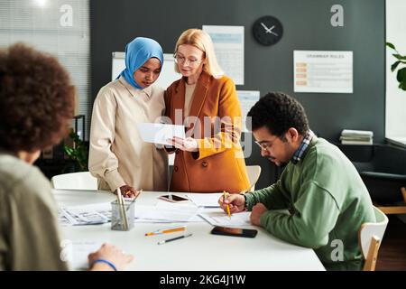 Professeur blond mûr montrant un document avec un test de grammaire anglaise à une étudiante musulmane en hijab tout en étant debout sur le lieu de travail Banque D'Images