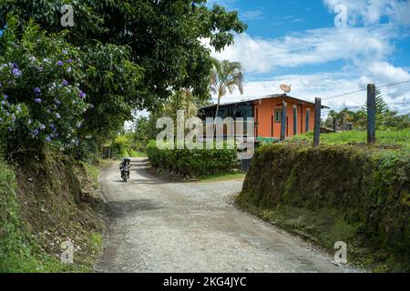 Filandia, Quindio, Colombie - 6 juin 2022: Une moto passant par une route de terre entourée d'un grand nombre d'arbres, de plantes et d'une maison d'orange Banque D'Images