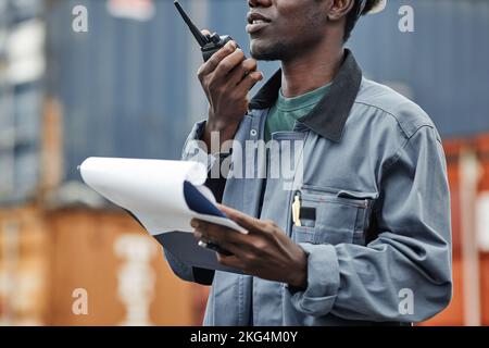 Prise de vue rognée d'un jeune homme noir utilisant un talkie-walkie tout en travaillant sur des quais d'expédition avec des conteneurs Banque D'Images