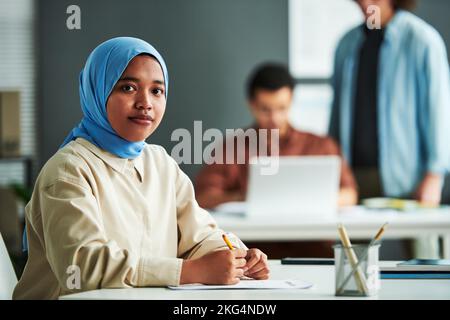 Jeune femme musulmane en hijab bleu regardant la caméra tout en vérifiant ou en effectuant un test de grammaire par le lieu de travail contre deux gars Banque D'Images