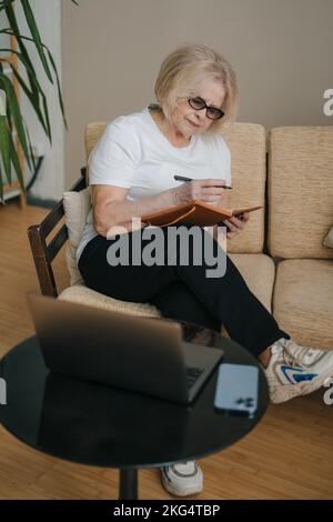 Portrait d'une femme âgée aux cheveux gris, assise sur un canapé, prenant des notes dans un bloc-notes à la maison avec un ordinateur portable ouvert. Communication Internet. Éducation Banque D'Images