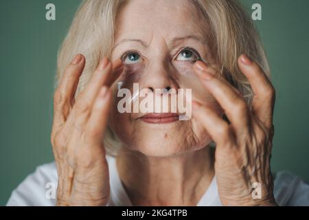 Gaie ridée femme sénior touchant le visage en appliquant délicatement des patchs hydratants de collagène sous les yeux. Concept anti vieillissement de la cosmétologie. Thérapie Banque D'Images