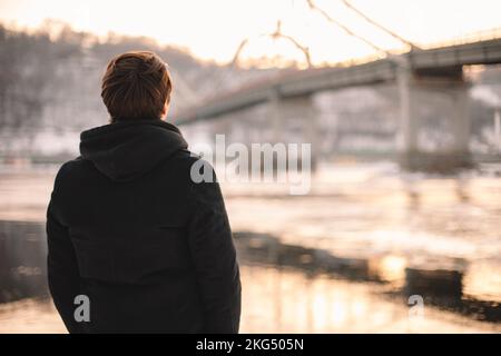 Vue arrière d'un jeune homme attentionné, debout au bord de la rivière en hiver Banque D'Images