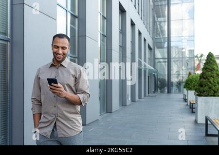 Jeune homme beau homme d'affaires hispanique, afro-américain marchant dans la rue près du centre de bureau. Il tient le téléphone dans sa main gauche, compose un numéro, vérifie le courrier, lit les nouvelles. Banque D'Images