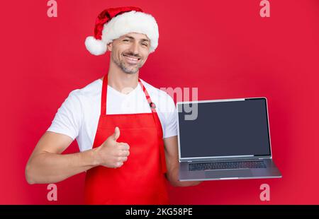 homme mature en chapeau de noël et tablier rouge présentant l'ordinateur portable. pouce vers le haut Banque D'Images