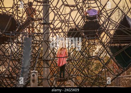 Les enfants jouant sur des cordes d'escalade au parc de jeu extérieur avec l'accent sur les petites filles en chemise rose et les cheveux longs blonds - après-midi - heure d'or Banque D'Images