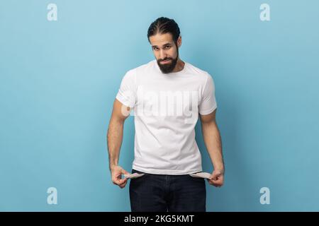 Portrait de l'homme avec la barbe portant T-shirt blanc montrant des poches vides et regardant frustré par les prêts et les dettes, n'a pas d'argent, sans emploi. Studio d'intérieur isolé sur fond bleu. Banque D'Images