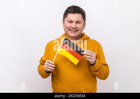 Satisfait positif homme d'âge moyen tenant le drapeau allemand dans sa main et regardant l'appareil photo avec un sourire crasseux, portant le sweat à capuche de style urbain. Studio d'intérieur isolé sur fond blanc. Banque D'Images