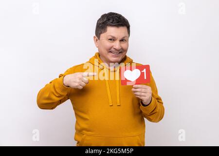 Blogs Internet. Portrait d'un homme joyeux pointant du cœur comme une icône, recommandant de cliquer sur le bouton des médias sociaux, portant un sweat à capuche de style urbain. Studio d'intérieur isolé sur fond blanc. Banque D'Images