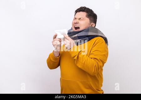 Homme enveloppé d'un foulard éternuant dans une serviette, nettoyant le nez et la toux, souffrant de symptômes de la grippe, fièvre, portant un sweat à capuche de style urbain. Studio d'intérieur isolé sur fond blanc. Banque D'Images