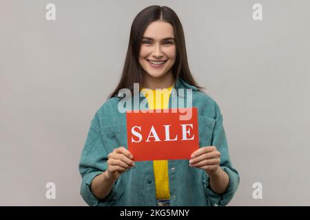 Portrait de jeune adulte heureuse souriante femme debout tenant une carte avec inscription à la vente, réductions énormes, portant une veste de style décontracté. Prise de vue en studio isolée sur fond gris. Banque D'Images