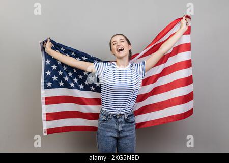 Portrait de jeune adulte gai belle femme portant un T-shirt rayé tenant le grand drapeau américain, exprimant le bonheur, criant heureusement, réjouissant. Prise de vue en studio isolée sur fond gris. Banque D'Images