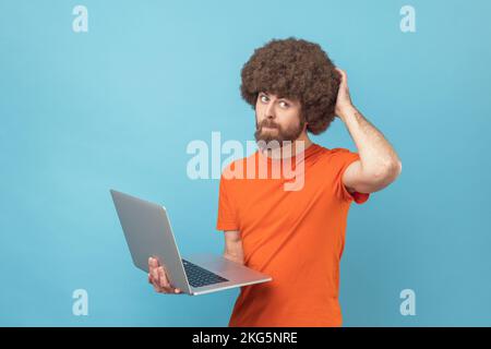 Portrait d'un homme avec une coiffure afro portant un T-shirt orange tenant un ordinateur portable et pensant à un nouveau projet, en grattant l'arrière de sa tête. Studio d'intérieur isolé sur fond bleu. Banque D'Images