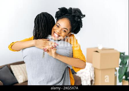 Excitée joyeuse femme afro-américaine avec des dreadlocks, embrassant son mari en se tenant dans le salon, heureuse femme montrant les clés de leur nouvelle propriété, elle regarde l'appareil photo, souriant Banque D'Images