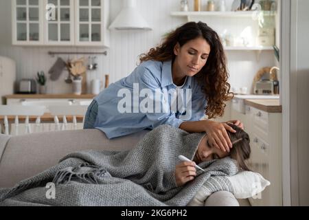 Gentille femme attentive met la main sur le front de la fille allongée sur le canapé souffrant de frissons Banque D'Images