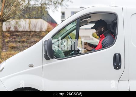 Courrier ethnique mâle à l'intérieur d'une camionnette de livraison blanche assise sur le siège du conducteur et woking sur son ordinateur portable. Temps d'automne. Concept de livraison de colis. Photo de haute qualité Banque D'Images