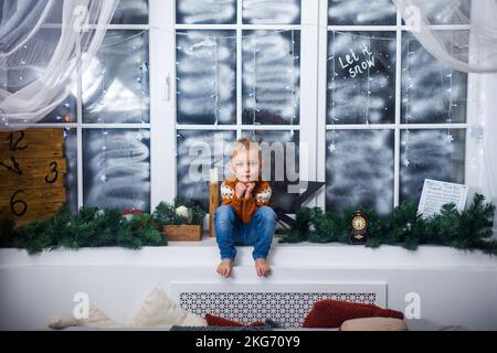 Garçon dans un chandail est assis sur le rebord de la fenêtre à la maison, en attendant des miracles, des cadeaux et Noël Banque D'Images