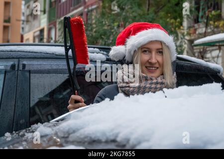 Une jeune femme dans un chapeau de Père Noël avec une brosse à neige est debout près de la voiture, va nettoyer sa voiture Banque D'Images