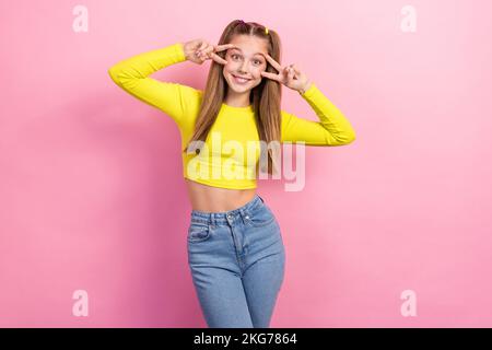 Portrait d'une fille optimiste avec des cheveux droits habillé jaune manches longues montrant le V-signe près des yeux isolés sur fond jaune Banque D'Images