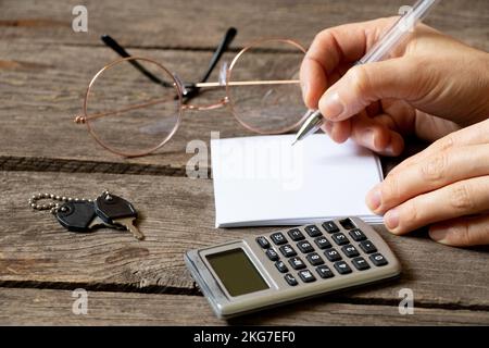 une table et la main d'une femme écrivent avec un stylo sur une feuille de papier à côté se trouve une calculatrice, des clés et des lunettes, un lieu de travail, un bureau Banque D'Images