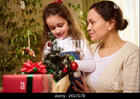 Une mère affectueuse et affectueuse qui embrasse sa adorable petite fille, qui pendre des balles de jouet sur un petit arbre de Noël écologique Banque D'Images