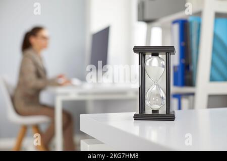 Le verre de sable au bureau sur un arrière-plan de bureau flou avec une femme travaillant sur un ordinateur. Banque D'Images