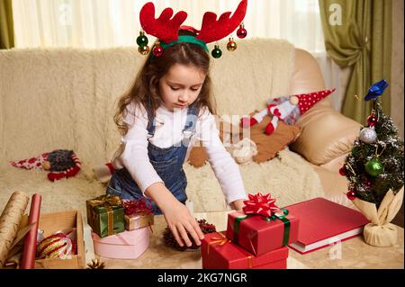 Adorable petite fille portant un cerceau de cerf, debout à table avec des cadeaux de Noël. Noël. Joyeuses vacances d'hiver Banque D'Images