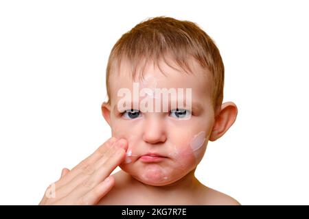 La mère sent la crème allergique sur le visage du bébé tout-petit, fond blanc studio. Portrait en gros plan d'un bébé mignon avec de la crème sur ses joues. Enfant âgé d'un an Banque D'Images