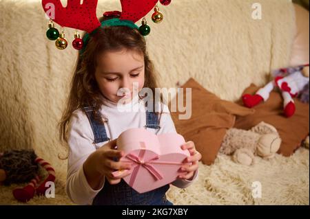 Charmante petite fille caucasienne portant un cerceau de cerf, tenant une boîte cadeau en forme de coeur rose. Lendemain de Noël. Noël Banque D'Images