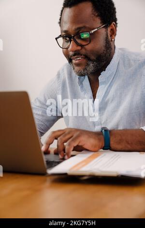 Homme barbu afro-américain travaillant sur un ordinateur portable tout en étant assis près du bureau à la maison Banque D'Images