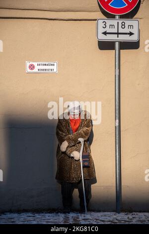 Femme âgée en vêtements d'hiver avec une béquille et le soleil qui lui brillent, penchée et reposant contre un mur sans panneau de route de stationnement, vertical Banque D'Images