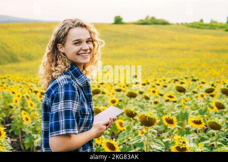 Souriante bonne femme agriculteur travaillant sur le terrain, en utilisant une application mobile sur une tablette numérique dans le champ de tournesol. sace libre pour le texte. Concept de Banque D'Images