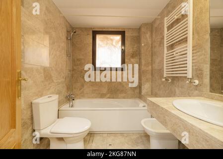 Salle de bains avec miroirs revêtus de marbre, sol en marbre crème, comptoir assorti, base en porcelaine et baignoire en résine blanche avec porte-serviettes chauffant Banque D'Images