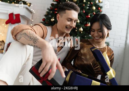 Joyeux couple interracial ouvrant Noël présent tout en étant assis sous le sapin décoré Banque D'Images