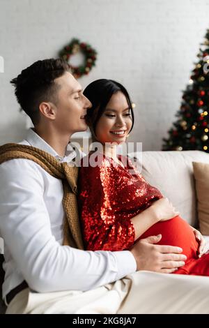 Femme asiatique enceinte et élégante souriant près de son mari embrassant son ventre dans le salon avec décoration de Noël Banque D'Images