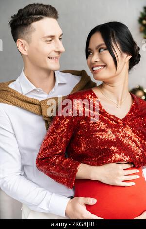 homme heureux et femme asiatique enceinte en vêtements de fête se souriant les uns les autres Banque D'Images