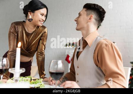 Couple interracial gai regardant l'un l'autre près de repas de fête et de verres de vin pendant Noël Banque D'Images