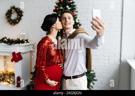 femme asiatique enceinte et élégante embrassant mari prenant selfie sur téléphone portable dans le salon décoré Banque D'Images