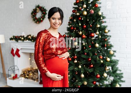 Gaie et enceinte asiatique femme en rouge élégant vêtements regardant l'appareil photo près de l'arbre de Noël décoré Banque D'Images