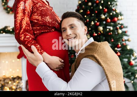 Un homme heureux s'est penché sur le ventre de sa femme enceinte dans un chemisier rouge brillant lors de la fête de Noël Banque D'Images