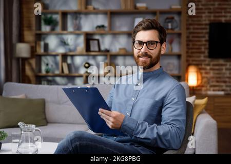 Un homme beau adulte s'assoit à la maison dans un bureau élégant. Psychologue, coach ou homme d'affaires regardant la caméra et souriant. Belles dents et Banque D'Images
