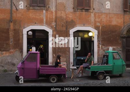 Rome, Italie: L'homme s'assoit à l'arrière d'un camion vert ouvert; parle à une femme qui se détend sur un petit camion rose. Véhicules garés à l'extérieur de Lettere Caffe à Trastevere. Banque D'Images