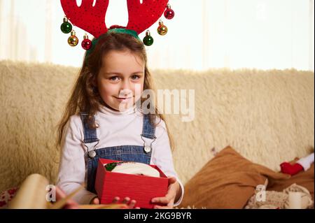 Une petite fille adorable portant un cerceau de cerf, sourit en regardant l'appareil photo tout en déballant une magnifique boîte cadeau rouge. Noël Banque D'Images