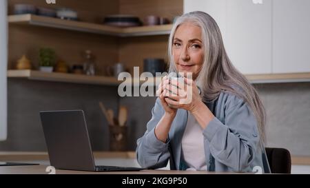 Femme d'affaires caucasienne femme au foyer senior indépendant femme mature assis à la cuisine à la maison tient une tasse de thé ou de café chaud dans les mains garde au chaud travaux sur Banque D'Images