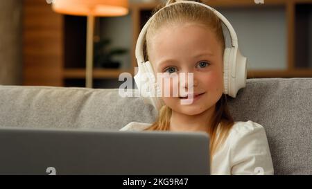 Adorable enfant heureux jolie fille dans les écouteurs s'asseoir à l'appartement écouter de la musique sur ordinateur portable jouer jeux d'ordinateur drôle petit enfant écofille montre tout-petit Banque D'Images