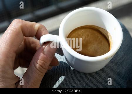 Gros plan de la main humaine avec une petite tasse de café espresso blanc et vide, en plein soleil. Moment de méditation. Frein à café. Banque D'Images