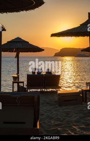 IOS, Grèce - 13 septembre 2022 : vue d'un couple allongé sur des chaises longues, boire du vin et profiter de l'incroyable coucher de soleil orange dans iOS Grèce Banque D'Images