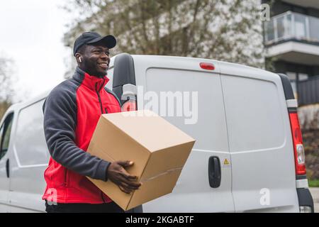 Concept de livraison d'achats en ligne. Positif souriant Noir homme à sa fin 20s tenant un gros paquet de carton. Prise de vue moyenne en extérieur. Camionnette de livraison blanche en arrière-plan. Photo de haute qualité Banque D'Images