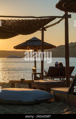 IOS, Grèce - 13 septembre 2022 : vue d'un couple allongé sur des chaises longues, boire du vin et profiter de l'incroyable coucher de soleil orange dans iOS Grèce Banque D'Images